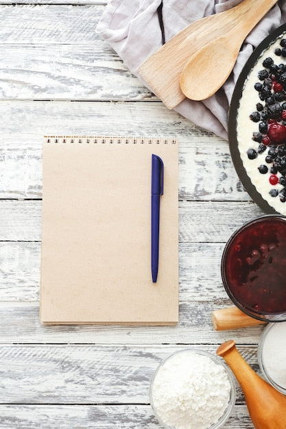 Leeres Notizbuch mit Stift auf Holztisch, daneben Beerenkuchen, Mehl, Marmelade und Holzlöffel
