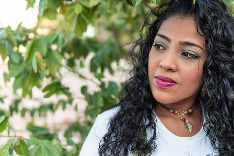 Frau mit lockigen Haaren und weißem Shirt steht draußen vor grünen Blättern