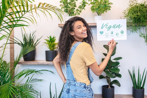 Junge Frau in Latzhose hält vegan Schild, umgeben von Zimmerpflanzen, lächelnd