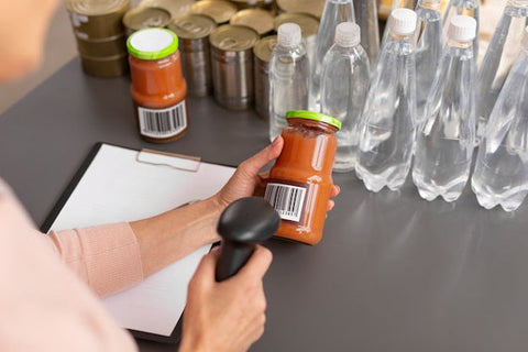 Mitarbeiter scannt Barcode auf Glas mit Tomatensauce, Wasserflaschen, Konservendosen, Lager