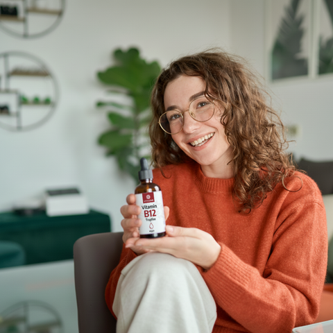 Frau mit Brille hält VITALOVE Vitamin B12 Tropfen Flasche im Wohnzimmer